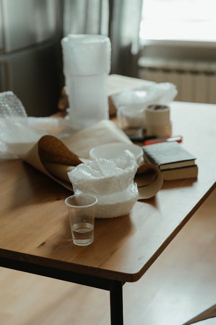 A wooden table inside a home shows various packing materials used for house moves, including multiple rolls of bubble wrap, crumpled packing paper, and a cardboard tube. A small, clear plastic cup is