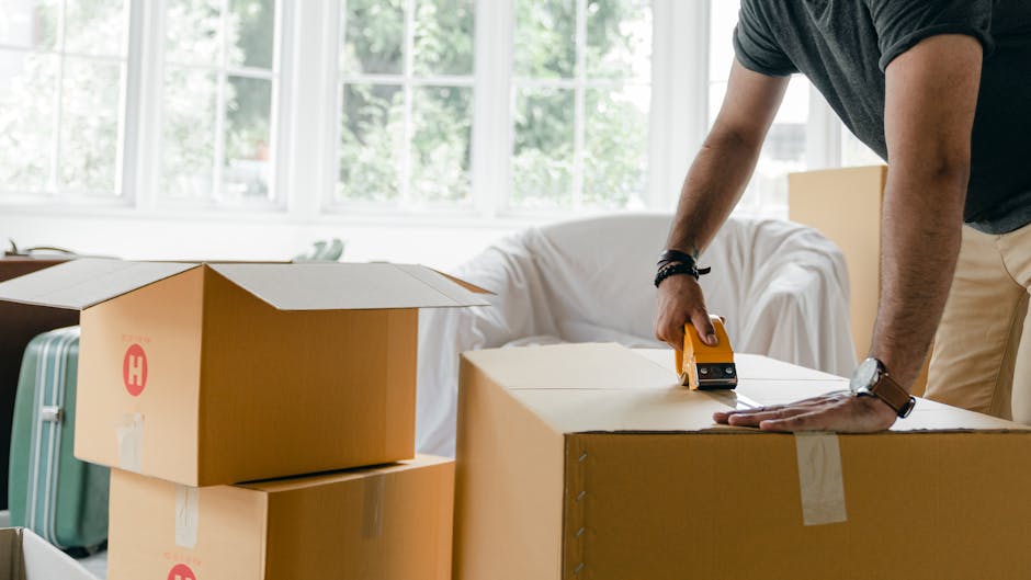 A wooden table inside a home shows various packing materials used for house moves, including multiple rolls of bubble wrap, crumpled packing paper, and a cardboard tube. A small, clear plastic cup is